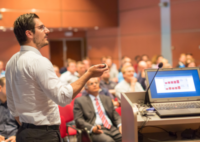 man giving a lecture next to a computer in front of a group of people